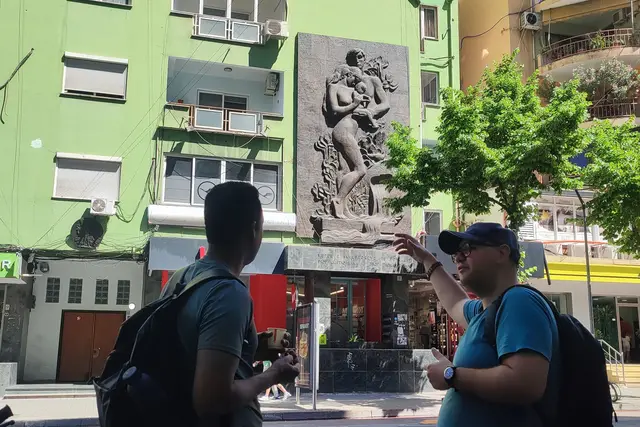 A guide pointing out a socialist-era relief to a guest on a Tirana street